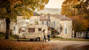 Un homme et une femme stationnent une petite caravane vintage près d'une cité historique française au début de l'automne, sous une lumière douce.