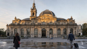 Vue immersive de la gare des Bénédictins à Limoges au petit matin, avec son dôme et son campanile éclairés par une lumière douce, ainsi que quelques voyageurs discrets.